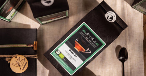Mehrere schwarze Papier-Teeverpackungen der Marke teealternative.de liegen auf einem Leinentuch. Im Vordergrund eine Packung "Grüner Tee" mit Tassen-Abbildung, daneben ein schwarzer Teelöffel.